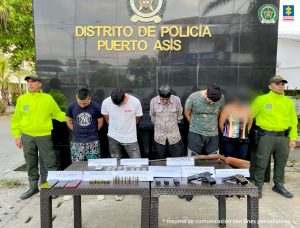 en la imagen se ven varias personas detenidas bajo custodia integrantes de la Policía Nacional. Frente a ellos una mesa con elementos incautados como armas de fuego, municiones y otros.