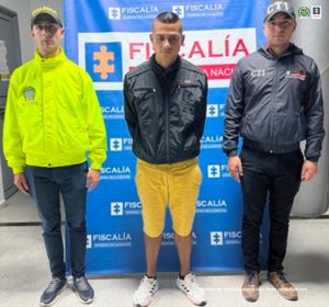 En la imagen se ve una persona detenida bajo custodia de un integrante del CTI de la Fiscalía y de la Policía nacional. Detrás suyo un backing de la Fiscalía.