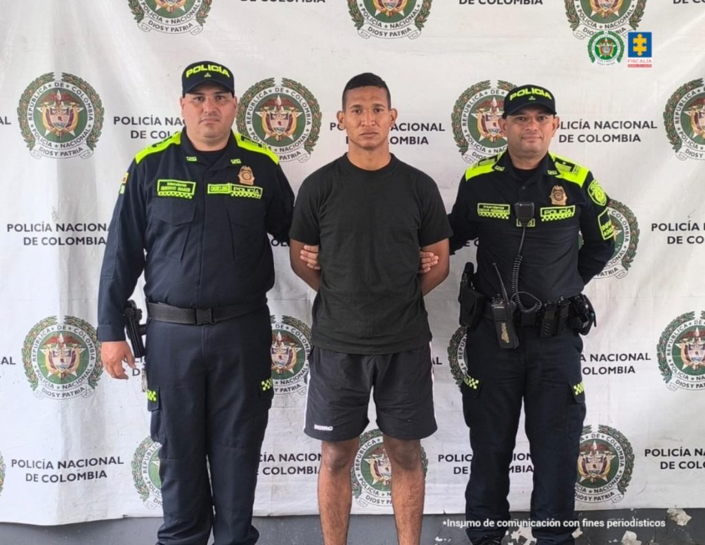 en la imagen se ve un hombre detenido bajo custodia de dos integrantes de la Policía Nacional. Detrás suyo un backing institucional.
