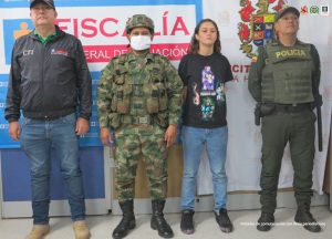 en la imagen se ve una mujer detenida bajo custodia de integrantes del CTI, la Policía, y el Ejército. Detrás suyo un backing institucional de la Fiscalía y otro del Ejército.