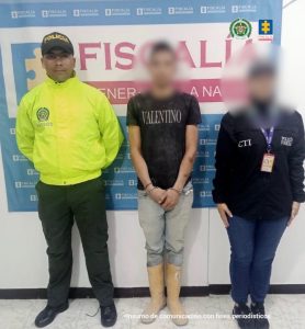 El capturado está de frente custodiado por investigador del CTI de la Fiscalía y Policía. Detrás banner de la Fiscalía.