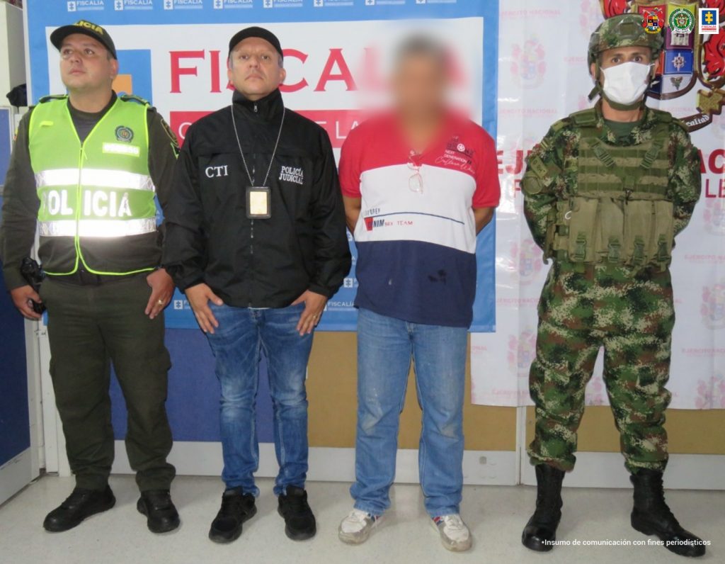 en la imagen se ve una persona detenida en custodia de un integrante de la Policía, Ejército y CTI. Detrás suyo un backing de la Fiscalía y del Ejército.