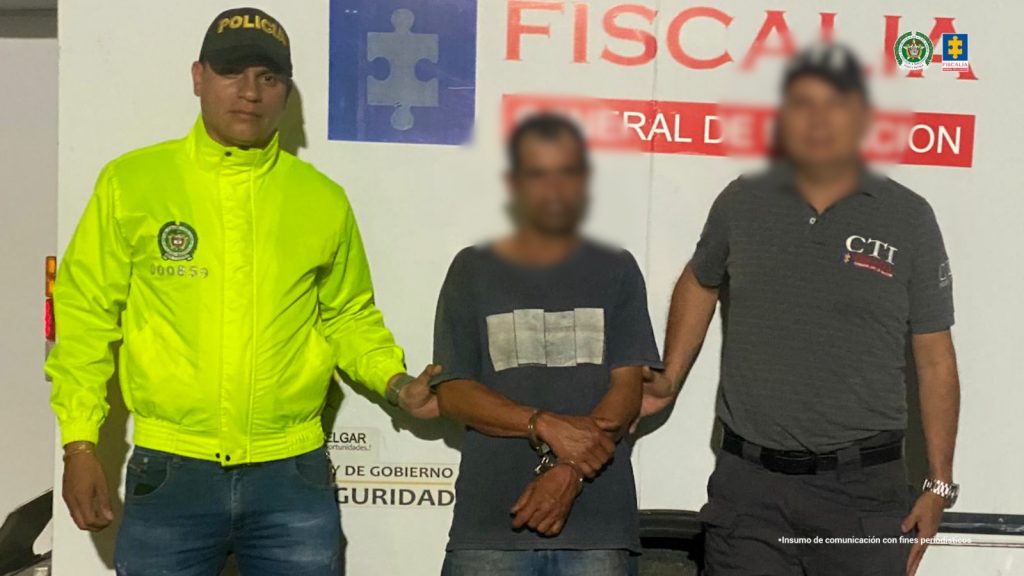 en la imagen se ve una persona detenida bajo custodia de un integrante de la Policía Nacional y de un investigador del CTI. Detrás suyo un backing institucional de la Fiscalía.