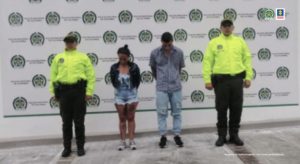 en la imagen se encuentran dos personas detenidas bajo custodia de dos integrantes de la Policía Nacional. Detrás suyo un backing de la institución.