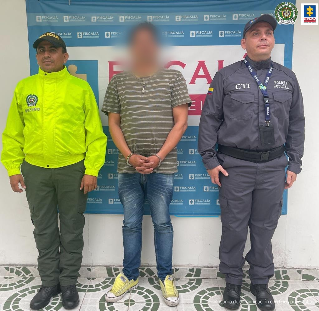 Capturado esposado en jean, camiseta y tennis, c ustodiado por investigador de CTI de Fiscalía y por un policía uniformados. Detrás banner de Ficalía.