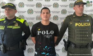 En la imagen se ve una persona detenida bajo custodia de dos uniformados de la Policía Nacional. Detrás suyo un backing institucional.