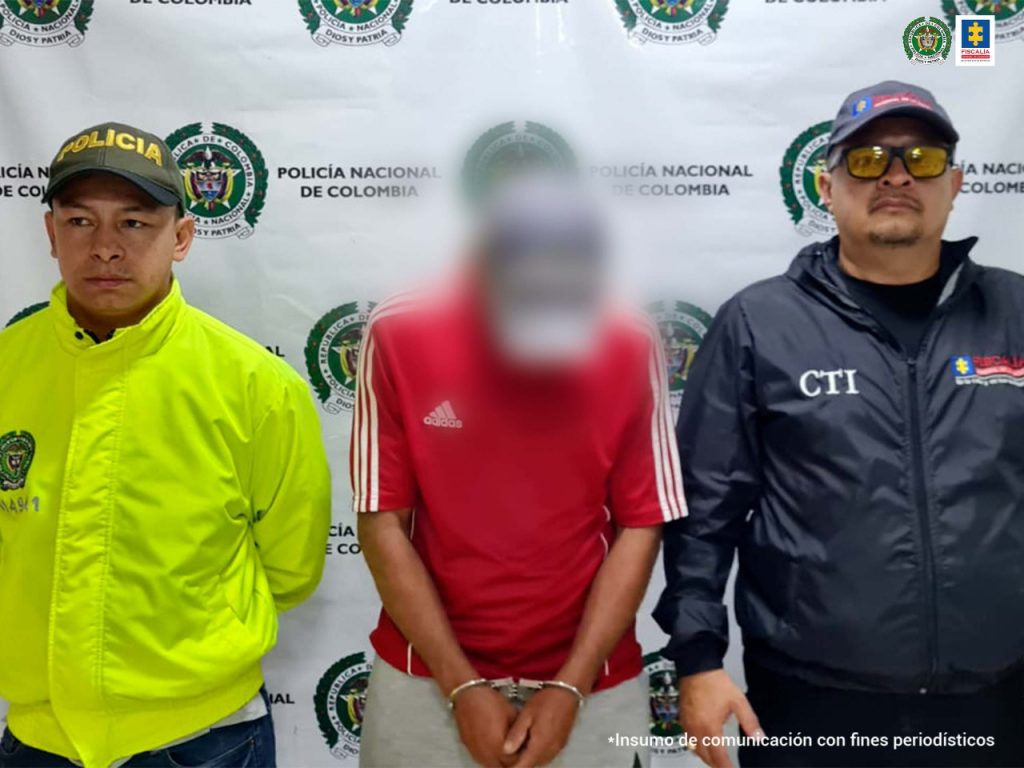 En la imagen se ve un hombre detenido el cual es custodiado por un investigador del CTI y de la Policía Nacional. Detrás suyo, un backing de esta última institución.