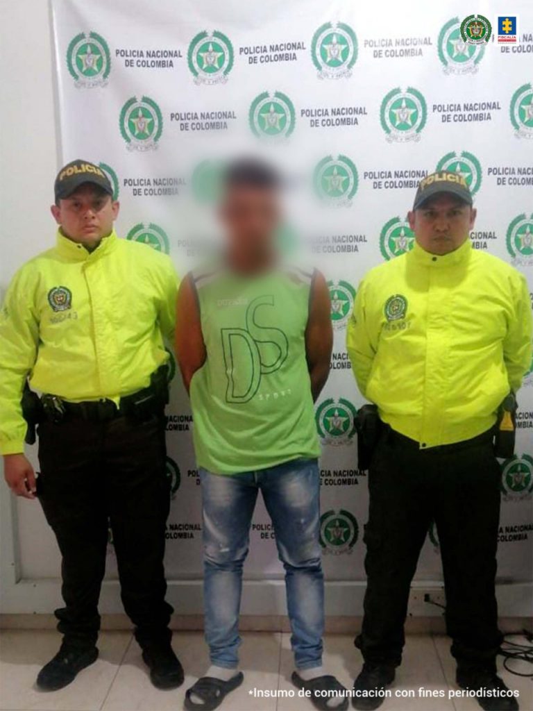 En la imagen un hombre detenido siendo custodiado por un funcionario del CTI de la Fiscalía. Detrás suyo un backing de la Policía Nacional y de la Fiscalía.