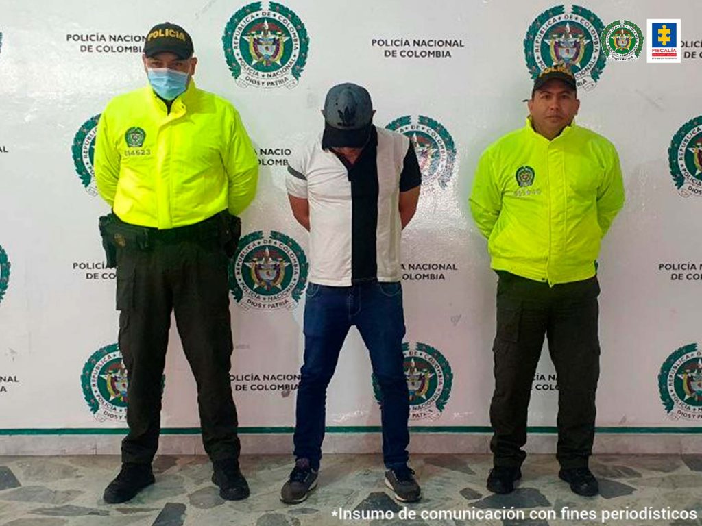 En la imagen se ve una persona detenida bajo custodia de dos uniformados de la Policía Nacional. Detrás suyo un backing de la institución.