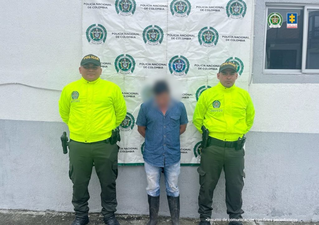 en la imagen se ve un hombre detenido bajo custodia de dos integrantes de la Policía Nacional. Detrás suyo un backing de la Policía.