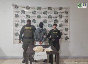 en la imagen se ve una persona detenida de espaldas bajo custodia de dos integrantes de la Policía Nacional. Detrás suyo un backing institucional.