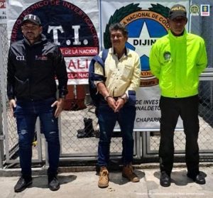 en la imagen se ve una persona detenida bajo cutodia de un investigador del CTI y de la Policía Nacional. DEtrás suyo un backing de la Fiscalía.