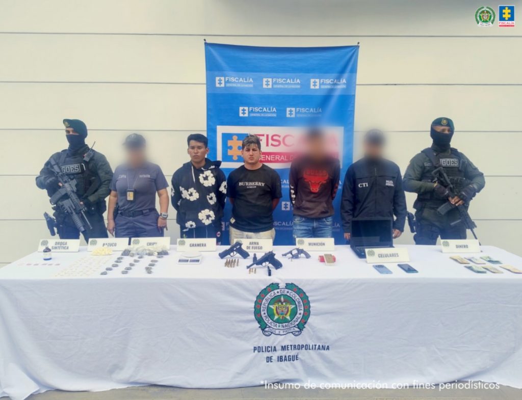 En la imagen se ven detenidas tres personas bajo custodia de investigadores del CTI e integrantes de la Policía Nacional. Delante suyo una mesa con elementos incautados, detrás, un backing institucional.