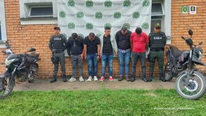En la imagen se ven cinco personas detenidas bajo custodia de dos integrantes de la Policía Nacional. Detrás suyo un backing institucional y a cada lado dos motocicletas.