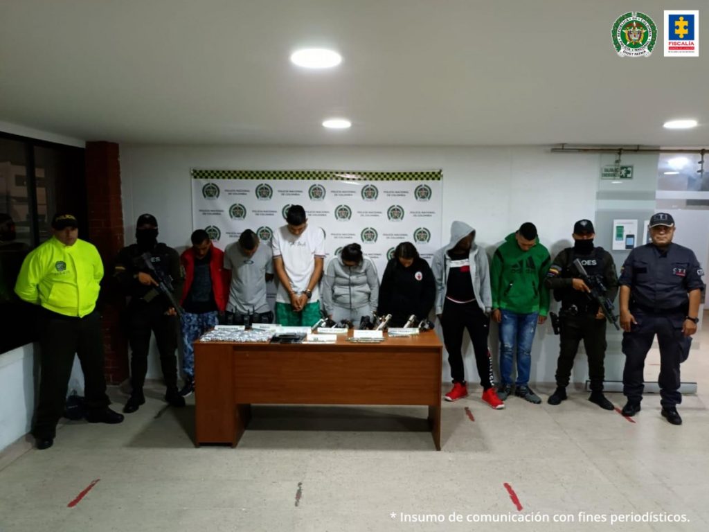 Se observan en fila site capturados. Al lado derecho un funcionario GOES y un funcionario CTI. Al lado izquierdo un funcionario GOES un funcionario Policía Nacional. Al fondo un pendón de Policía Nacional. Al frente una mesa con el material incautado en los allanamientos.