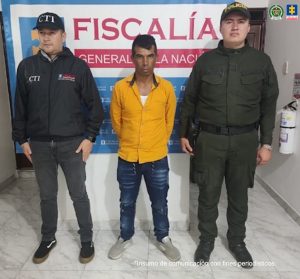  en la imagen se ve una persona detenida bajo custodia de un investigador del CTI y de la Policía. Detrás suyo un backing de la Fiscalía.