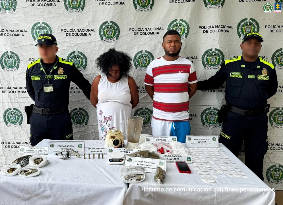 En la imagen está una mujer y un hombre morenos, están de pie, de frente con los brazos atrás esposados. La mujer es de contextura mediana con abultamiento en su abdomen tiene el cabello largo crespo negro y tiene un vestido blanco. El hombre tiene puesta una camiseta blanca con rayas rojas y pantaloneta azul con blanco. Se encuentran custodiado por dos uniformados de la Policía Nacional. 