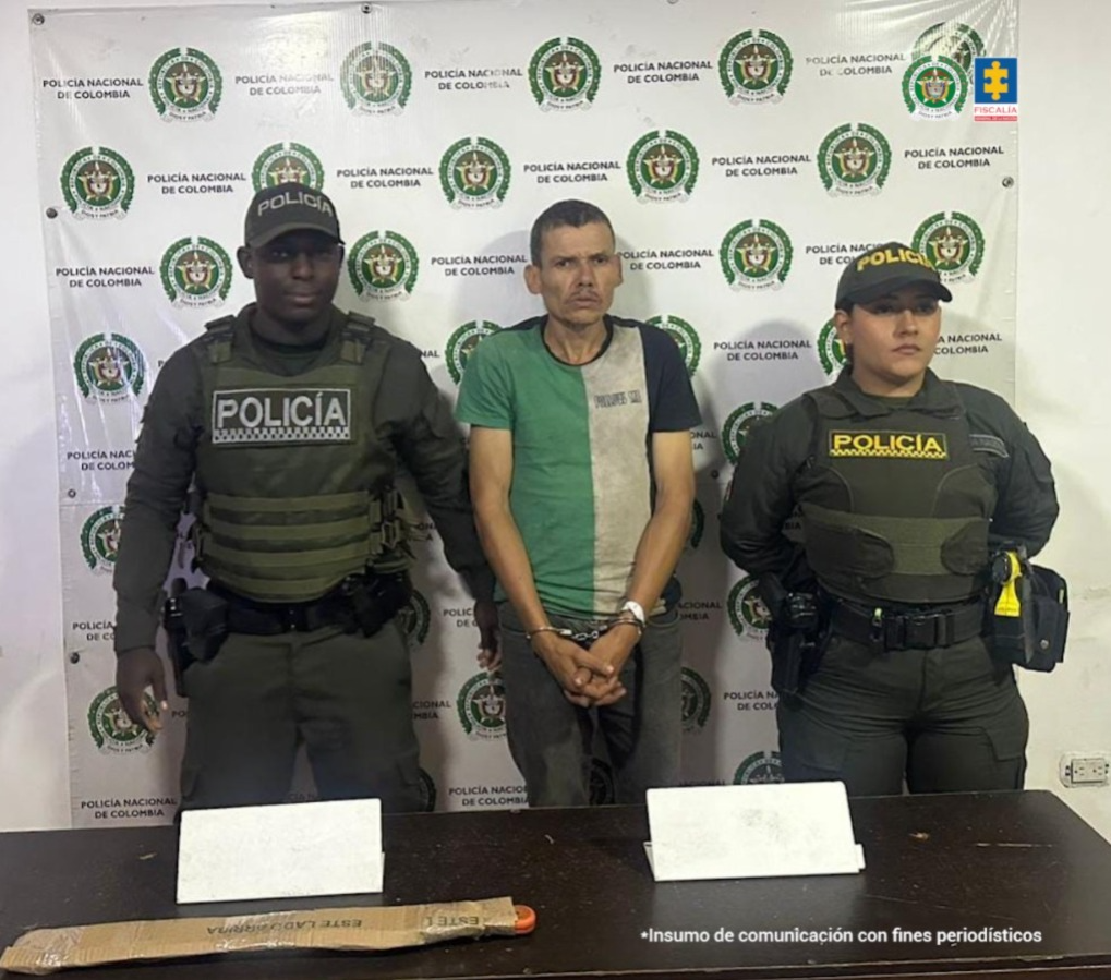 En la fotografía aparece de pie, con los brazos adelante y esposado Jesús Antonio Sanz Blanco. Viste con camiseta con franja verde en el lado derecho; franja gris en el izquierdo y manga de color negro en el brazo izquierdo; asimismo, pantalón de color gris oscuro. En sus extremos hay uniformados de la Policía Nacional. Delante de ellos hay una superficie marrón y sobre ella hay un contenedor de cartón alargado. Detrás de ellos hay un pendón institucional de la Policía.