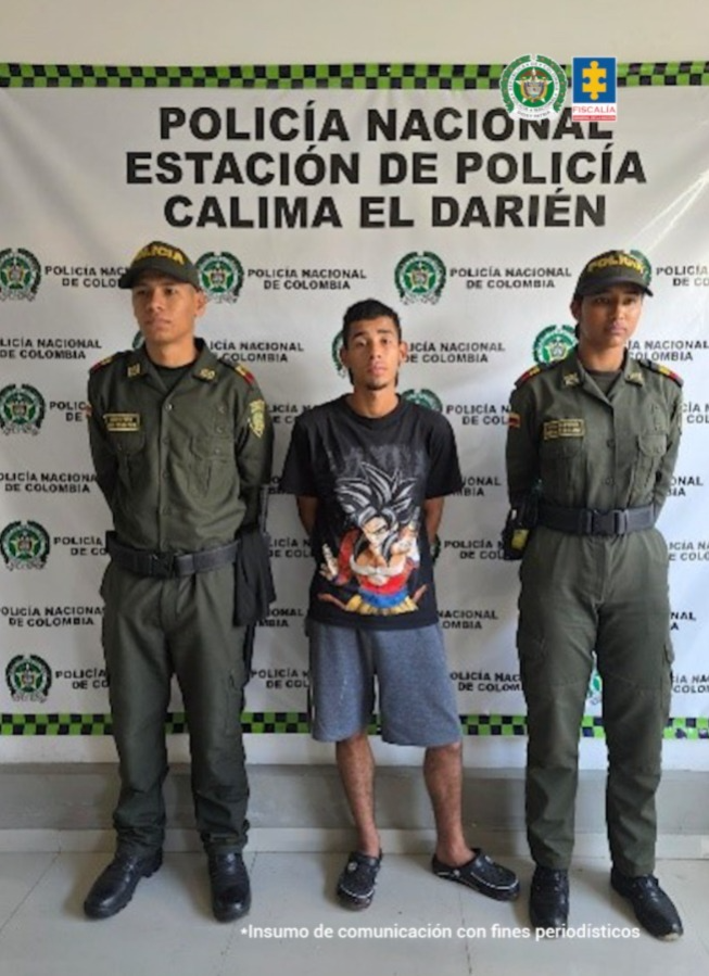 En la fotografía se aprecia de pie y con los brazos atrás a Jhon Jairo Usma Benítez. Viste con camiseta de color negro, mangas cortas y estampado grande de colores en la parte frontal; bermuda de color gris y pantuflas negras. En sus extremos hay uniformados de la Policía Nacional. Detrás de ellos hay un pendón institucional de la Policía.
