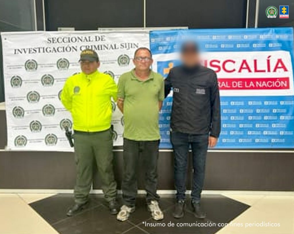 Se visualiza al capturado junto a un uniformado de la Policía Nacional y un investigador del CTI. Detrás de ellos los banners que identifican a la Policía Nacional y a la Fiscalía General de la Nación.