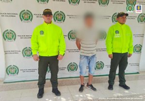 en la imagen un hombre detenido bajo custodia de la Policía, detrás suyo un backing institucional. 