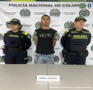 El capturado se observa de frente y a cada costado un funcionario de la Policía Nacional. Atrás un pendón de Policía Nacional