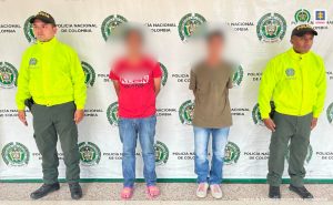 en la imagen se ven dos personas detenidas bajo custodia de dos integrantes de la Policía Nacional. Detrás suyo un backing institucional.  