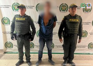 En la imagen se ve una persona detenida bajo custodia de dos integrantes de la Policía Nacional. Detrás suyo un backing institucional.  