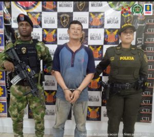 en la imagen se ve una persona detenida bajo custodia de dos integrantes de la Policía Nacional. Detrás suyo un backing institucional.