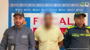 en la imagen se ve una persona detenida bajo custodia de un integrante del CTI de la Fiscalía y otro de la Policía. Detrás suyo un backing de la Fiscalía.