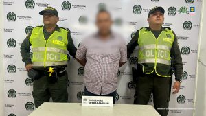 en la imagen se ve una persona detenida bajo custodia de dos integrantes de la Policía Nacional. Detrás suyo un backing institucional.