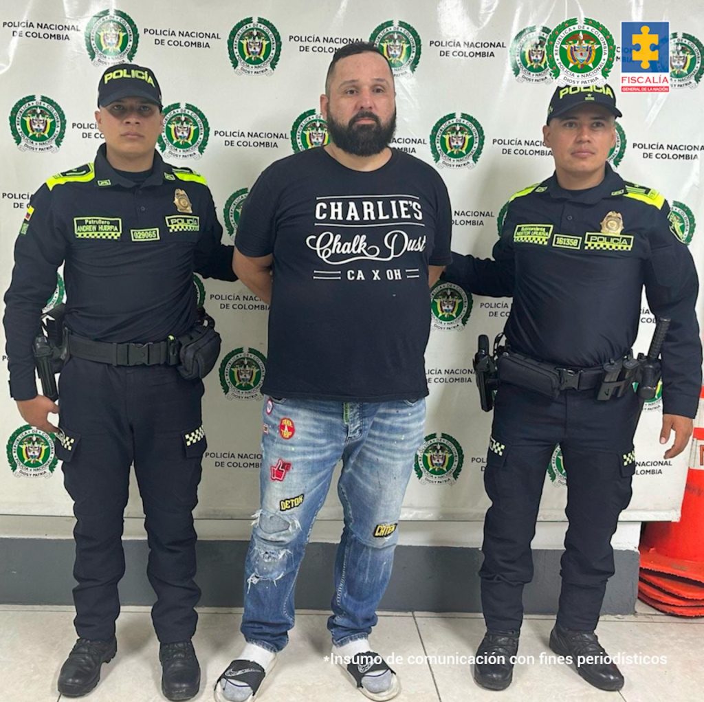 en la imagen se ve un hombre detenido bajo custodia de dos integrantes de la Policía Nacional. Detrás suyo un backing institucional.