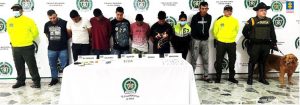 Los imputados se encuentra en custodia por dos uniformados de la Policía Nacional. Detrás se observa un backing de la Policía y se observa la mesa con el material y sustancias de alucinógenos incautados.