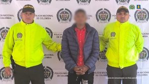 En la imagen se ve una persona de tenida bajo custodia de dos integrantes de la Policía NAcional. Detrás suyo un backing institucional.