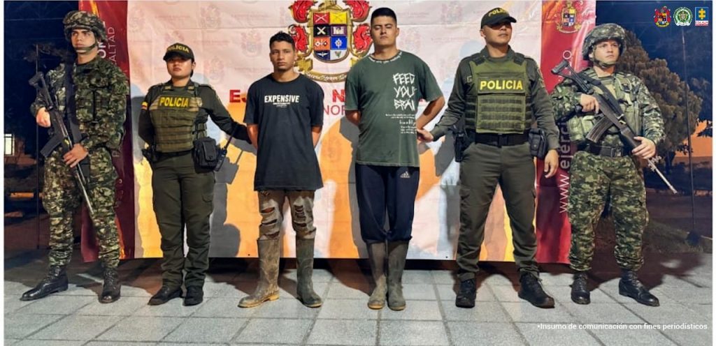 Dos hombres esposados, de pie en el centro, están custodiados por cuatro uniformados: dos soldados armados y dos policías. Todos posan frente a un pendón institucional con logos del Ejército, la Policía y la Fiscalía. Los dos detenidos visten ropa informal y botas de caucho.