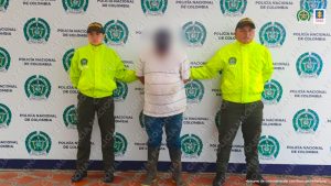 Se observa a un capturado con el rostro cubierto por un efecto difuminados en el centro y a cada costado un funcionario de Policía Nacional. Al Fondo pendón de Policía Nacional