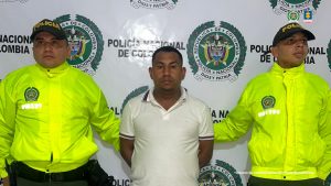 El capturado aparece en el centro de la imagen y en cada uno de sus costados hay miembros de la Policía. En la parte trasera se ve un banner de la Policía. 
