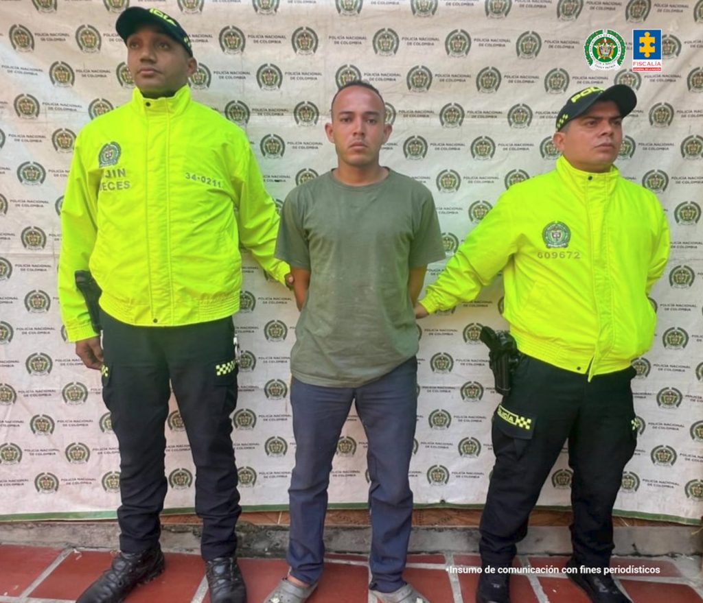 En la fotografía aparece una persona capturada, acompañado de dos uniformados de la Policía Nacional. En la parte posterior un banner con logos de la entidad. 