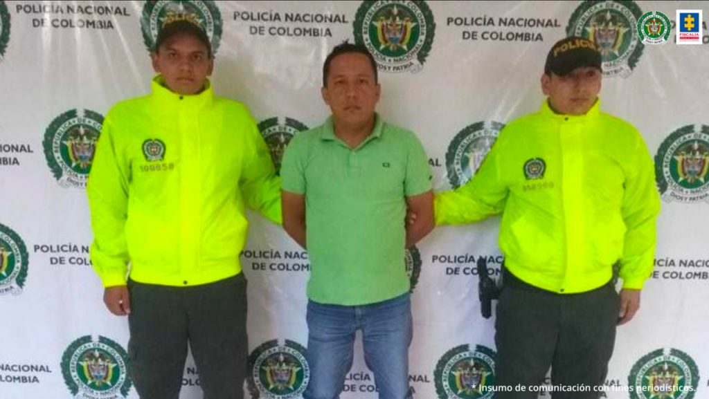 En la fotografía aparece un capturado, junto a personal de Policía Nacional. En la parte posterior de la imagen se ven banners de Policía Nacional y la Fiscalía General de la Nación.   