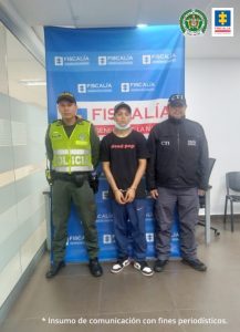 capturado con gorra de frente esposado manos adelante, custodiado por investigador del CTI de la Fiscalía y un policía. Detrás banner de la Fiscalía.
