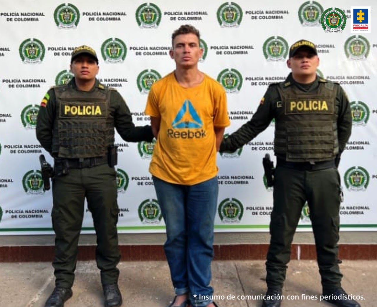 En la foto se ve un hombre con camiseta y jean de frente, custodiado por dos uniformados de la Policía Nacional y detrás de ellos un banner de esa institución.