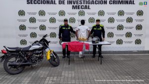 En la fotografía aparece de espaldas a la cámara, con los brazos atrás y esposado, Donald Stiven Riascos Gamboa. Viste con un camisa de color blanco con estampado amarillo en el borde de abajo y pantón blanco. Usa muletas y su pierna izquierda se encuentra vendada. En sus extremos hay uniformados de la Policía Nacional. En la parte delantera de hay una pesa y sobre se aprecia una chaqueta de color rojo y un casco. En el extremo izquierdo de la fotografía hay una motocicleta. En la parte posterior hay un pendón institucional de la Policía.