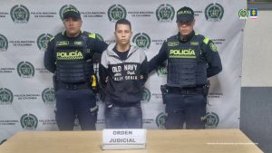 En la imagen se ve una persona detenida bajo custodia de dos uniformados de la Policía Nacional. Detrás suyo un backing institucional.   