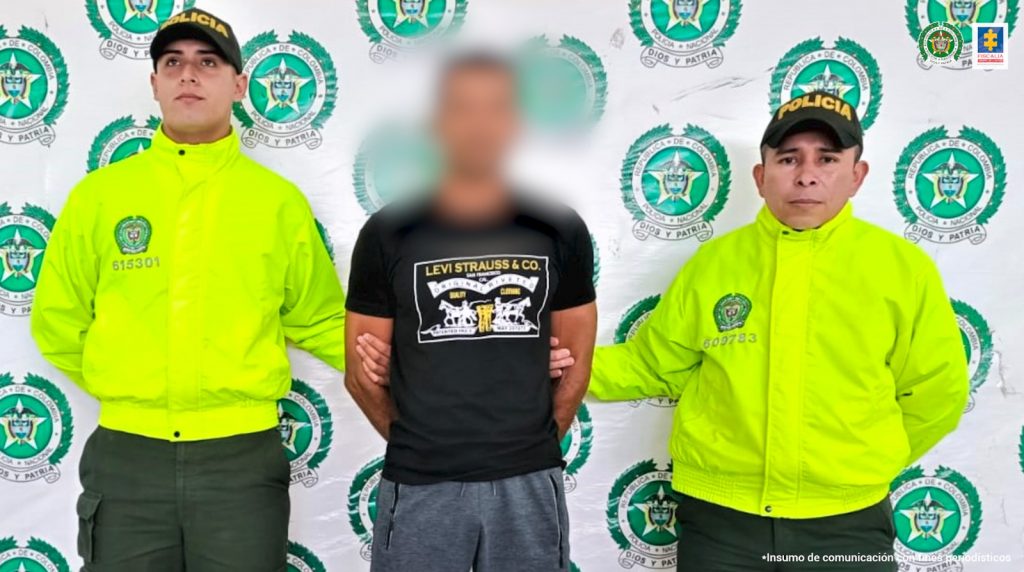 El capturado está esposado con sus manos a la espalda y custodiado por dos uniformados de la Policía Nacional.