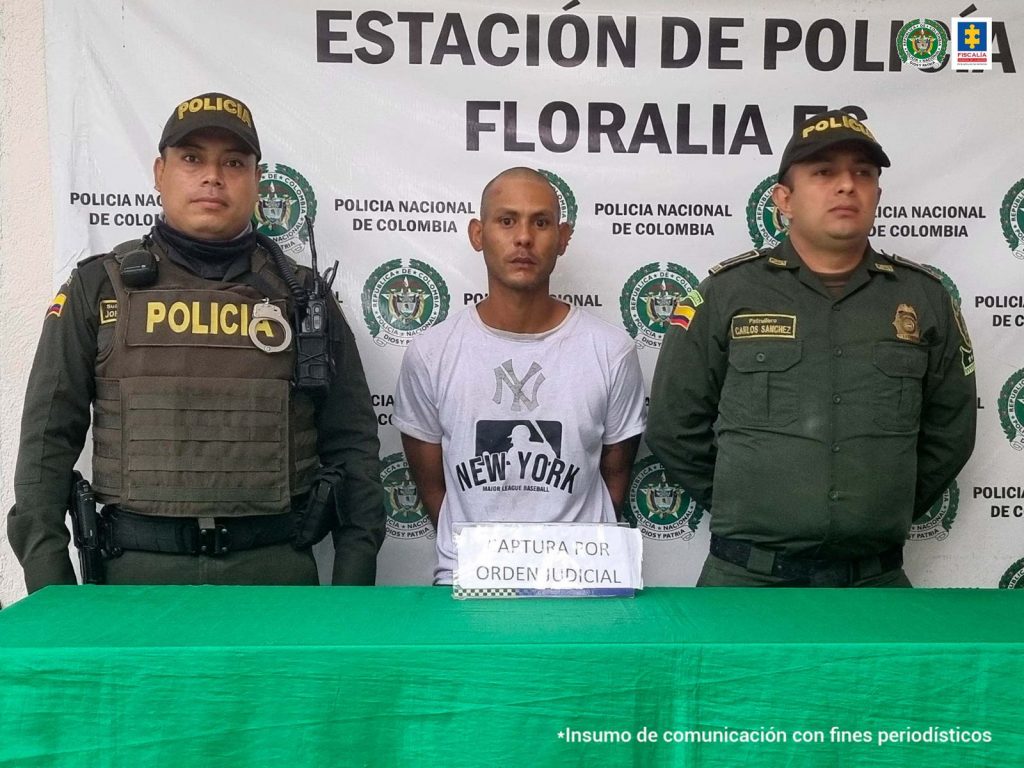 En la fotografía se aprecia a Alberto Alexander Griman Durand, alias Chamo. Viste una camiseta blanca de mangas cortas, con estampado a la altura del pecho con las iniciales NY y el nombre New York. A lado y lado de él hay uniformados de la Policía Nacional. Delante de ellos hay una mesa con mantel verde y sobre ella un letrero que dice: ‘Captura con orden judicial’. Detrás de ellos hay un pendón institucional de la Policía.