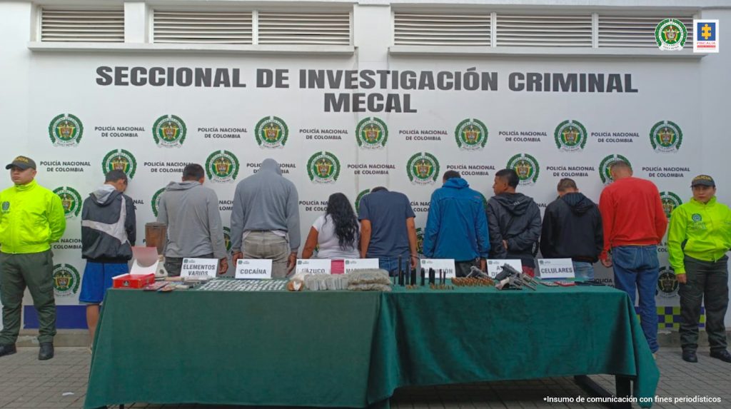 En la fotografía se aprecia a los presuntos integrantes de ‘Los del Filo’. Se encuentran de pie y de espaldas a la cámara. A lado y lado de ellos hay uniformados de la Policía Nacional. Se aprecia una mesa con mantel verde y sobre ella varios elementos y letreros. En la parte posterior hay un pendón institucional de la Policía.