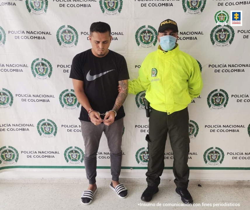 En fotografía se observan dos personas uno el capturado y el otro un integrante de la Policía Nacional. En la parte posterior se ve un backing con escudos de la Policía Nacional.