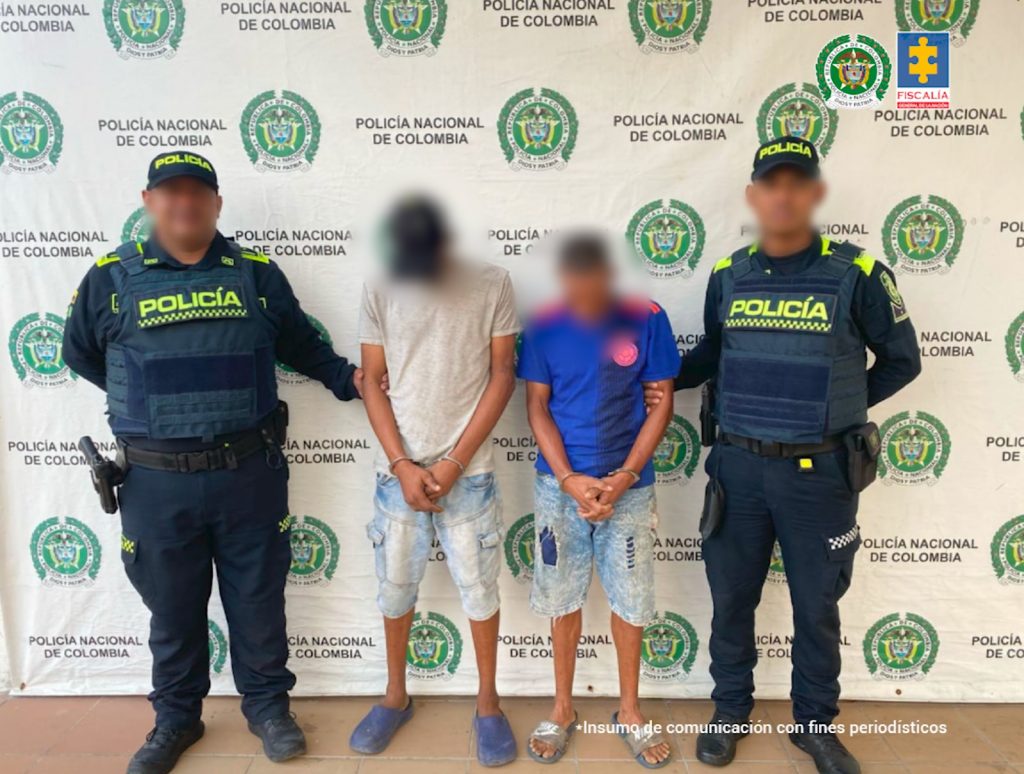 En la imagen están dos hombres de pie, uno alto y el otro de baja estatura, ambos delgados, con los brazos adelante esposados, vestidos con camisetas y bermudas y custodiado por dos uniformados de la Policía Nacional. 