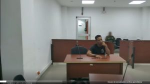 En la fotografía está un hombre moreno, con cabellos crespos negros, se encuentra sentando frente a un escritorio de madera con un micrófono, vestido de camiseta manga corta negra en la sala de audiencia donde se le procesó.
