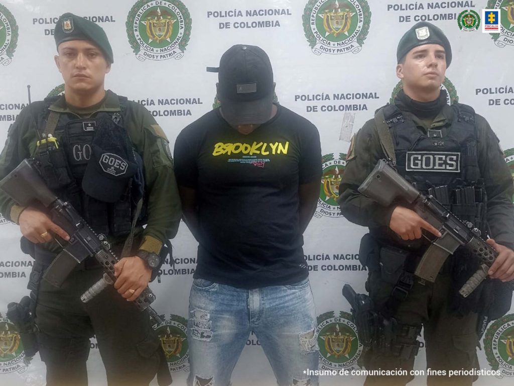 En la fotografía se aprecia a Moisés Rodallego López, de pie, con los brazos atrás y la cabeza agachada. Viste buzo oscuro con estampado a la altura del pecho que dice ‘Brooklyn’ en amarillo; jean azul desgastado y gorra oscura. A lado y lado de él hay uniformados de la Policía Nacional. Delante de ellos se aprecia parte de una superficie de color madera clara y sobre ella un letrero que dice: ‘Orden judicial’. Detrás de ellos hay un pendón institucional de la Policía.
